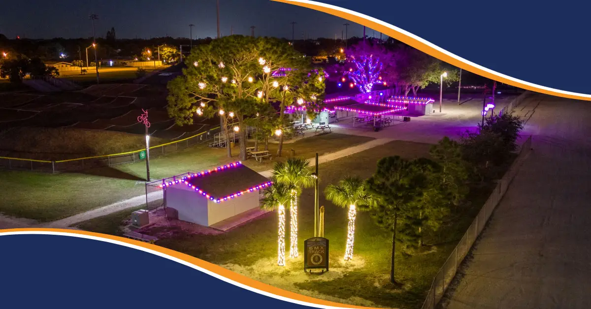 Commercial holiday lighting installation featuring colorful purple and pink roofline lights, illuminated trees, and festive outdoor lighting at night.